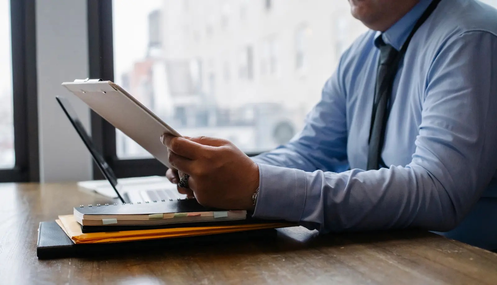 A man in a shirt and tie holds a clipboard with documents over a desk, next to a window
