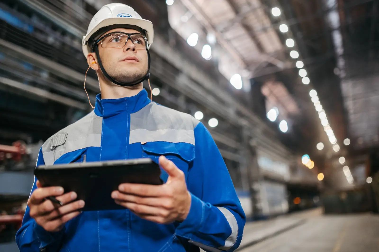 A worker in a white hard hat and blue coveralls holds a tablet in an industrial building