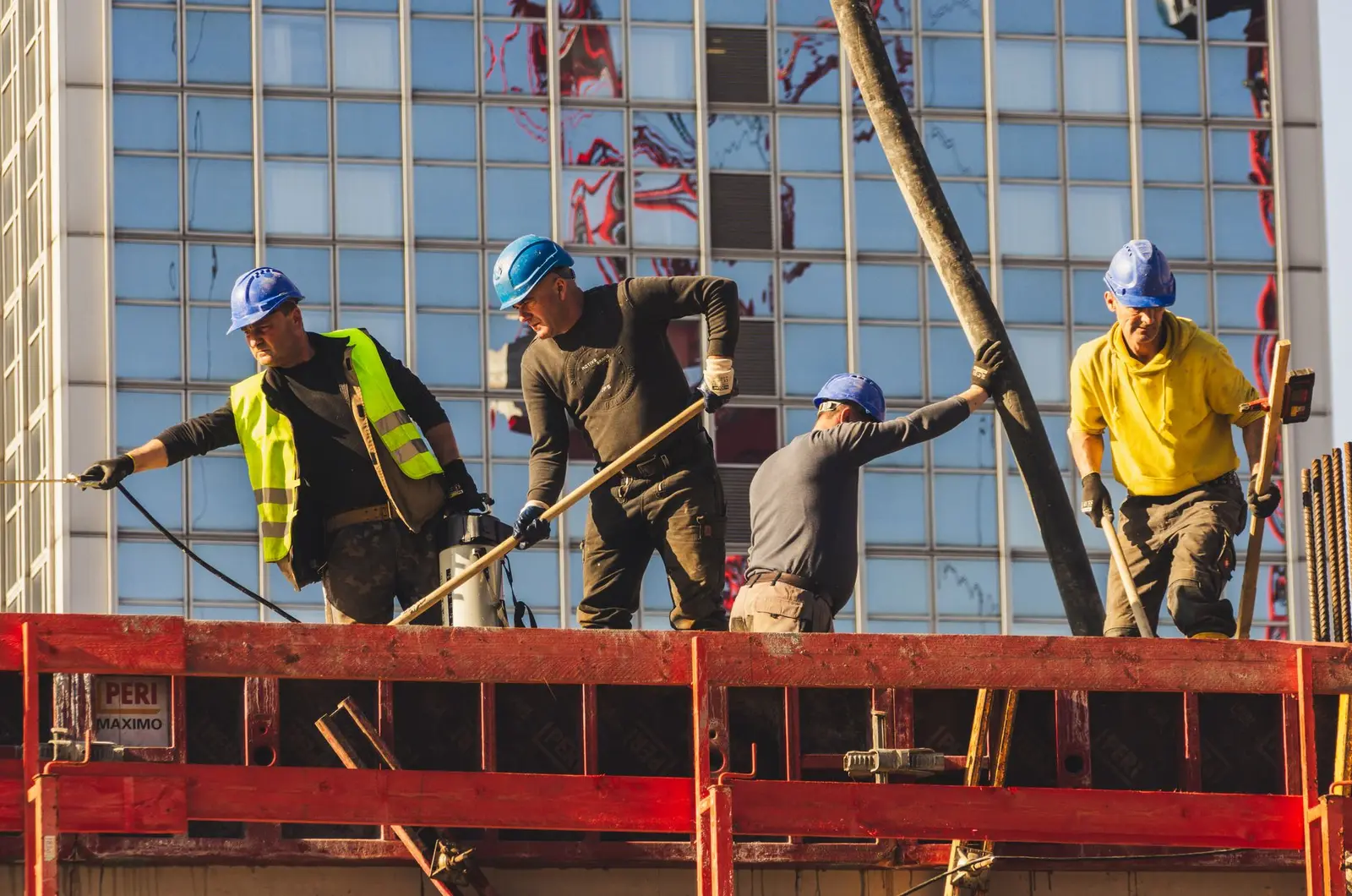 Four workers in hard hats on an elevated construction platform in front of a glass facade; one wears a reflective vest