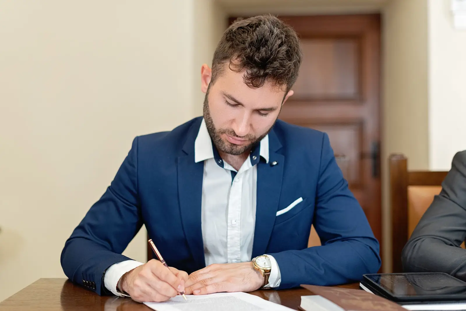 A man in a blue suit signs a document at a desk
