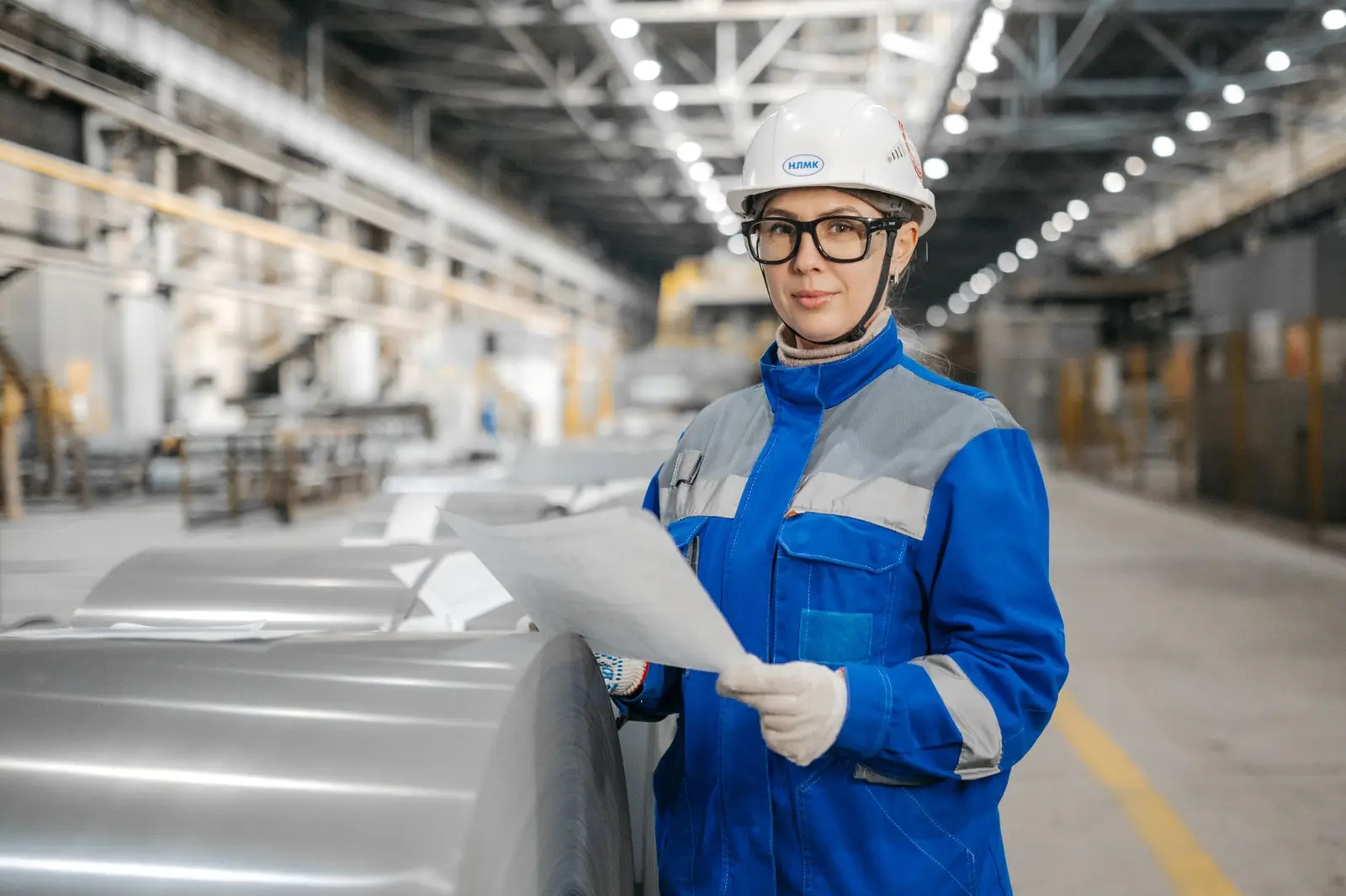 A worker in a white hard hat, safety glasses, and blue coveralls holds a document in an industrial plant